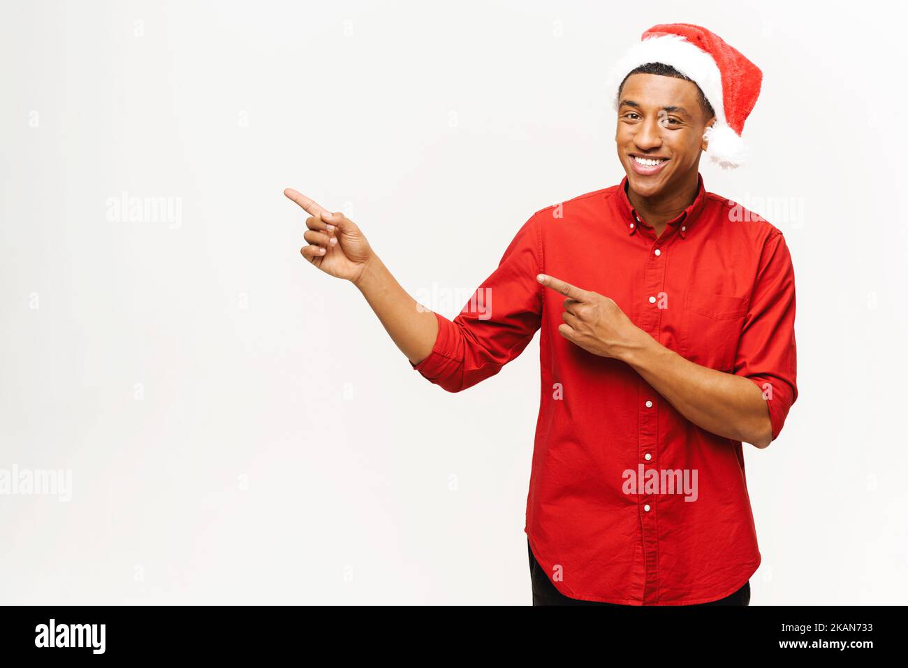 Incredible promotion. Youn gmultiracial man in santa hat points finger aside at empty copy space isolated on yellow, african-american guy draws attention to New Year advertising, holiday sales Stock Photo