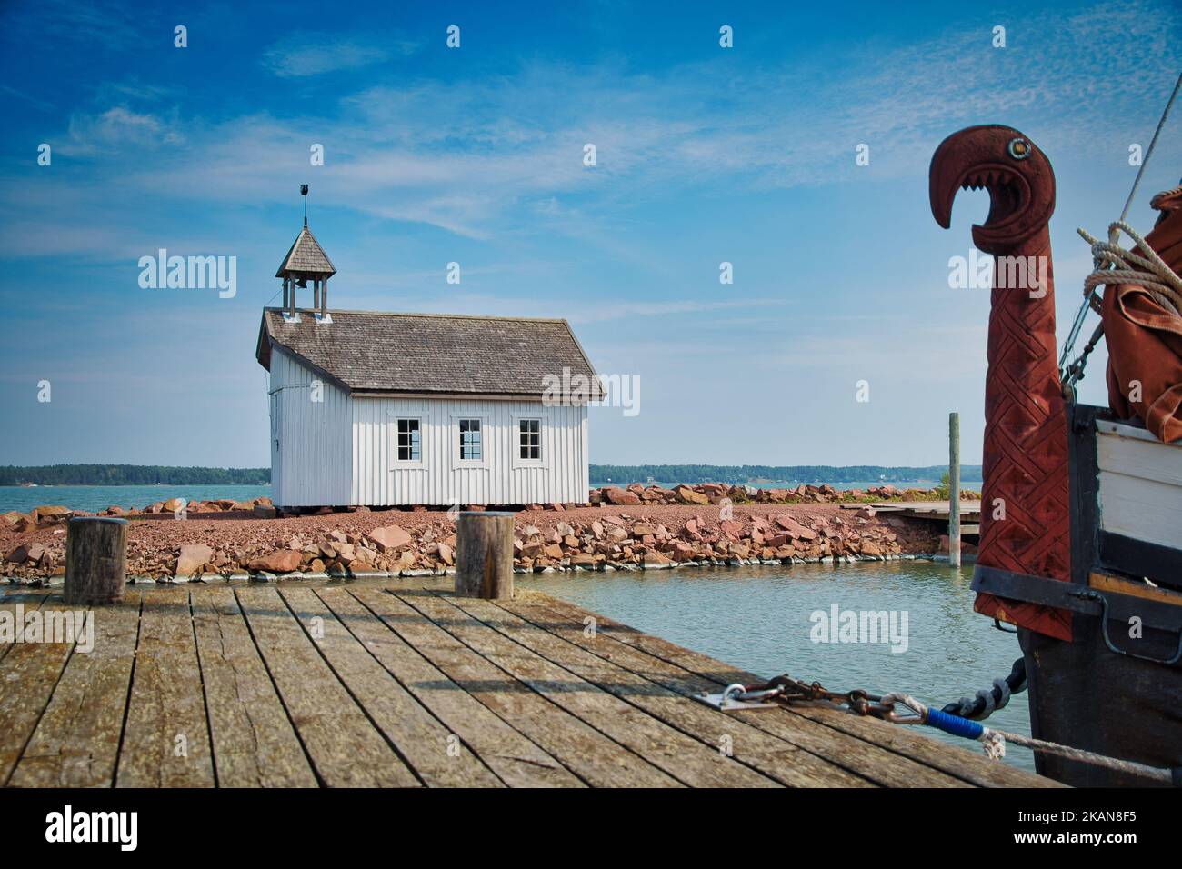A small wooden church on an island with a viking longship at the docks with a dragon's head on the prow Stock Photo