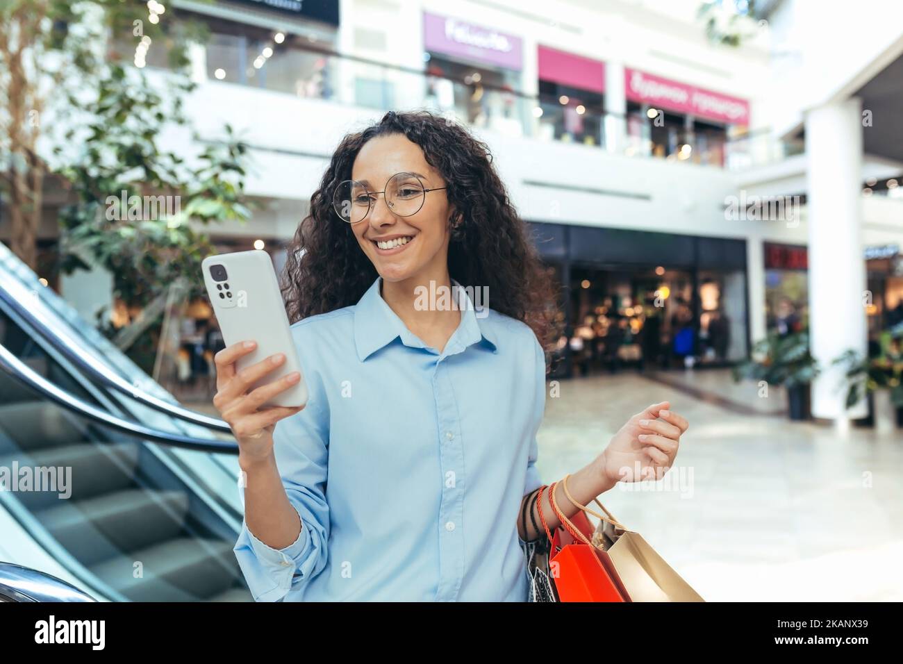 Happy hispanic woman in supermarket shopping, reading online message, holding colorful shopping bags and smartphone. Stock Photo