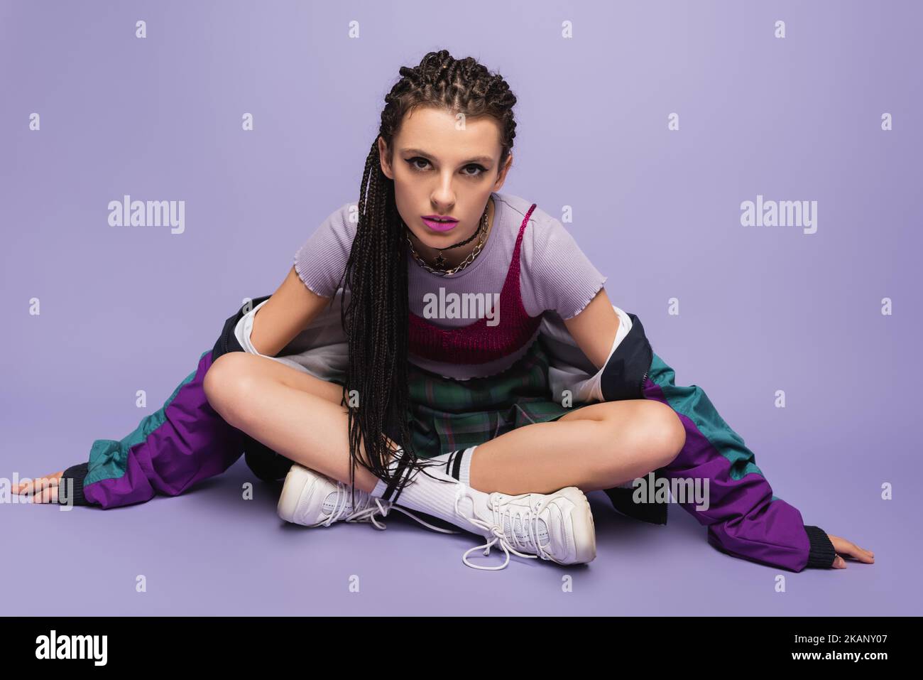 woman with braids sitting in casual nineties attire with crossed legs and looking at camera on purple background,stock image Stock Photo