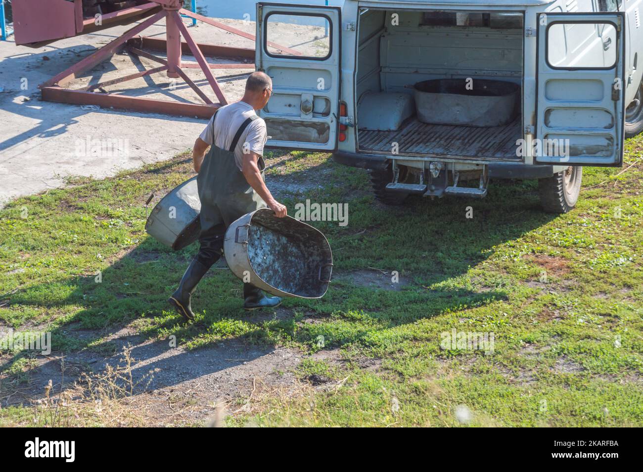 Fsherman in overall and rubber boots carries iron basins to car Stock Photo