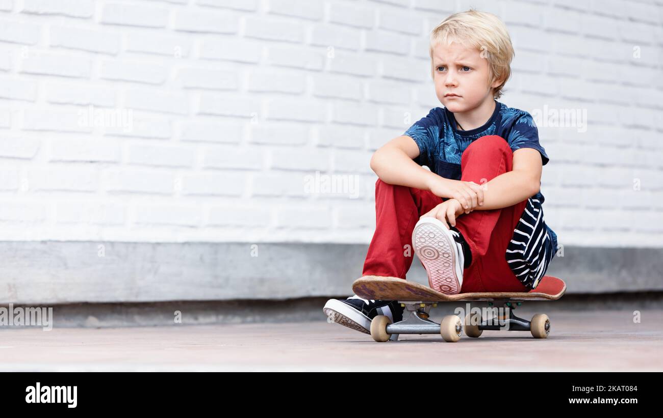 Looking unhappy and upset skater sit on skateboard before children training class in skate park. Active family lifestyle, outdoor activities Stock Photo