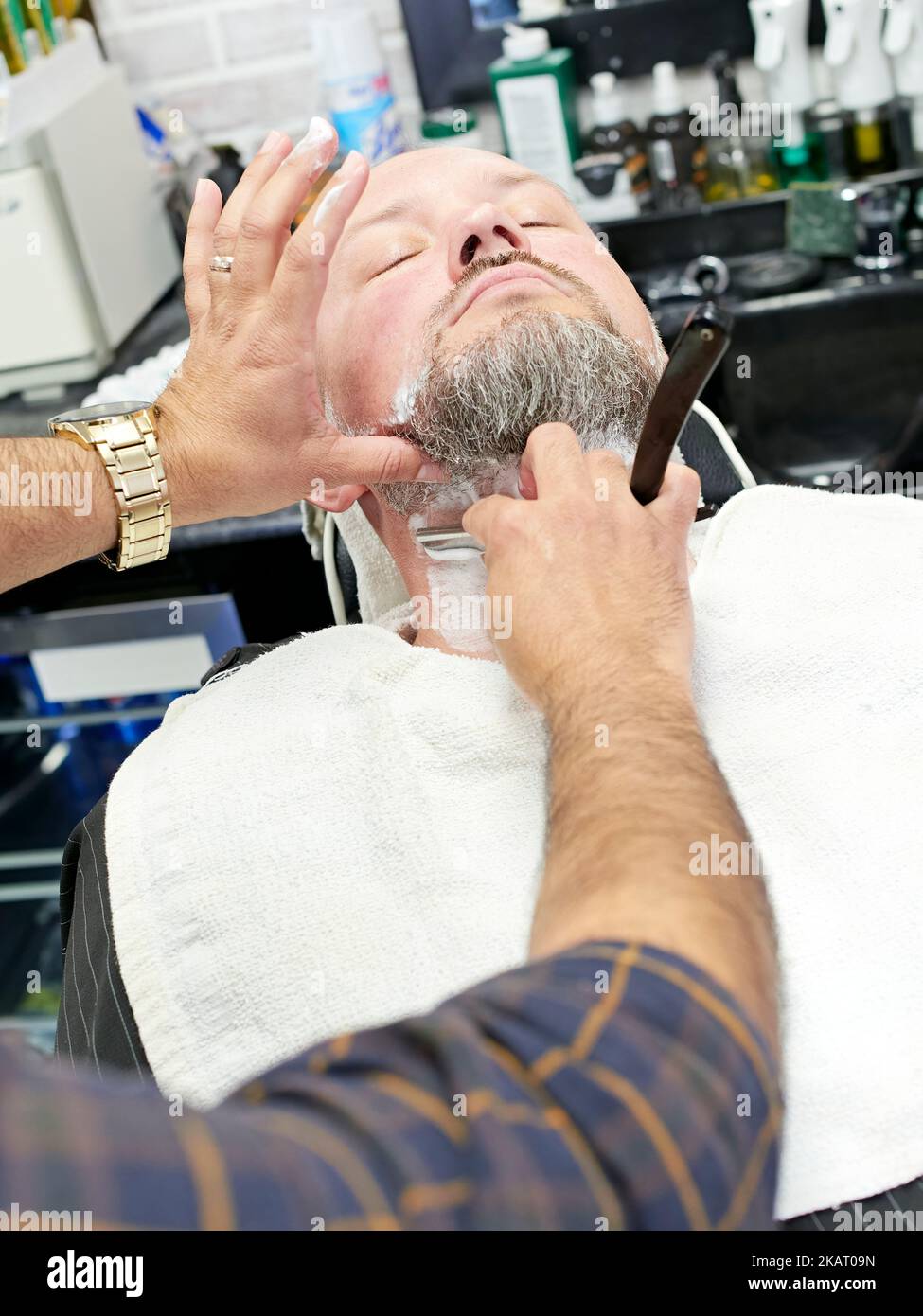 Man being shaved by a barber with an old fashioned straight razor while leaning back in a barber's chair at a barber shop. Stock Photo