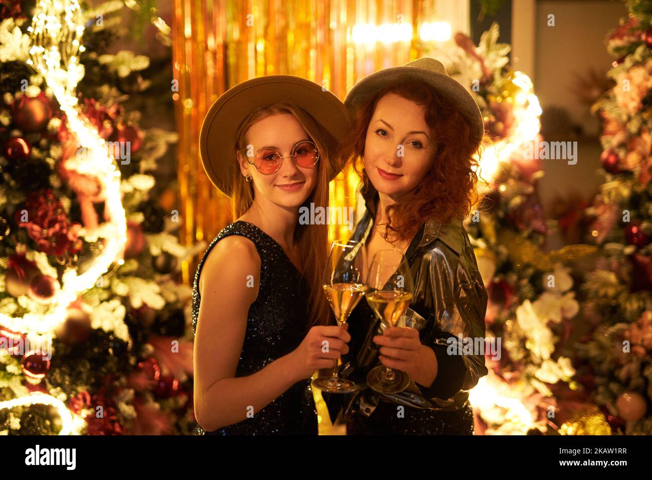 Happy and fun emotion during Christmas party. Caucasian mom and daughter talking with lights background. Females in hats holding glasses of sparkling wine during New Year's eve. High quality image Stock Photo