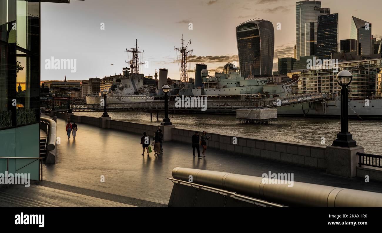 Couples walk along the promenade on the banks of the river Thames London walkie-talkie and the city in the background of HMS Belfast in the foreground the Thames London Stock Photo