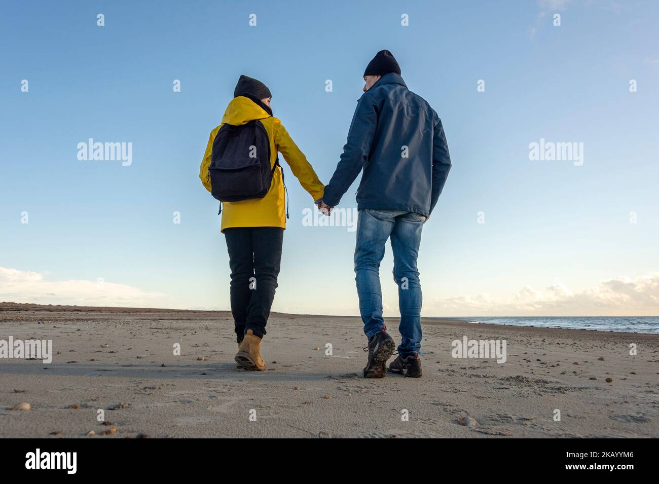 Rear view of a mature couple walking at beach holding hands, backpack and yellow coat, fall. Stock Photo