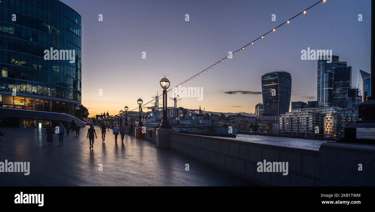 Close to Tower Bridge walkway with pedestrians on the Southbank looking across to the city the Thames and HMS Belfast in the foreground Stock Photo