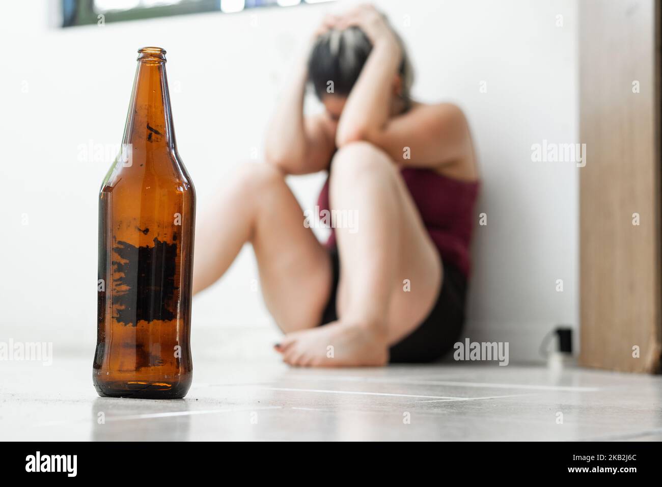 close-up of an empty beer bottle, placed on the ground, in the background a woman sitting on the ground in fetal position, crying because of domestic Stock Photo
