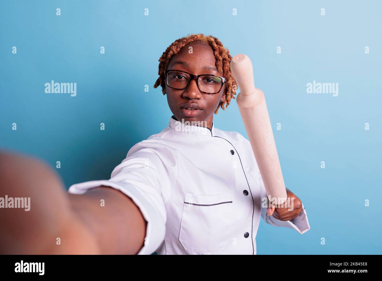 Angry female dinning hall subordinate threatening via video call. Annoyed african american chef intimidating with rolling pin. Self portrait of angry cooker in uniform. Stock Photo