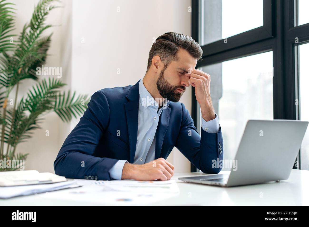 Frustrated overworked caucasian businessman, entrepreneur, product manager, programmer, sits at a desk in the office, massaging the bridge of his nose with his eyes closed, exhausted from long work Stock Photo