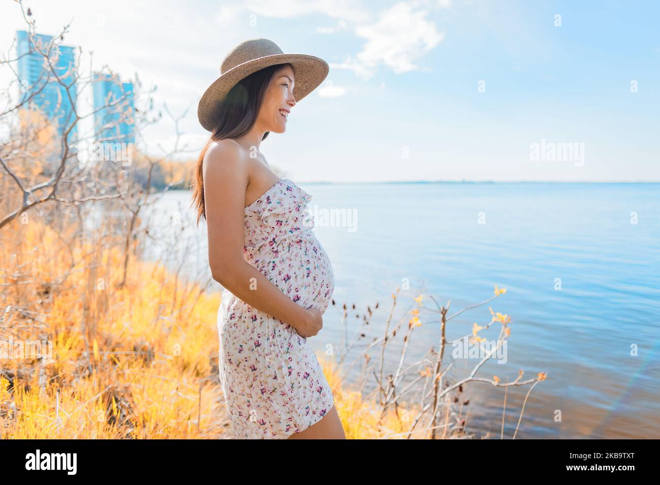 Pregnant Asian woman walking in city park by the lake wearing sun hat during spring nature walk relaxing lookig at landscape. Happy pregnancy outdoor Stock Photo