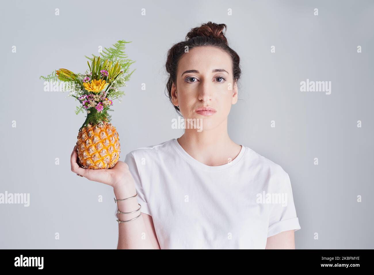 I like to decorate my fruit with flowers before I eat it. Studio shot of a beautiful young woman holding a pineapple against a grey background. Stock Photo