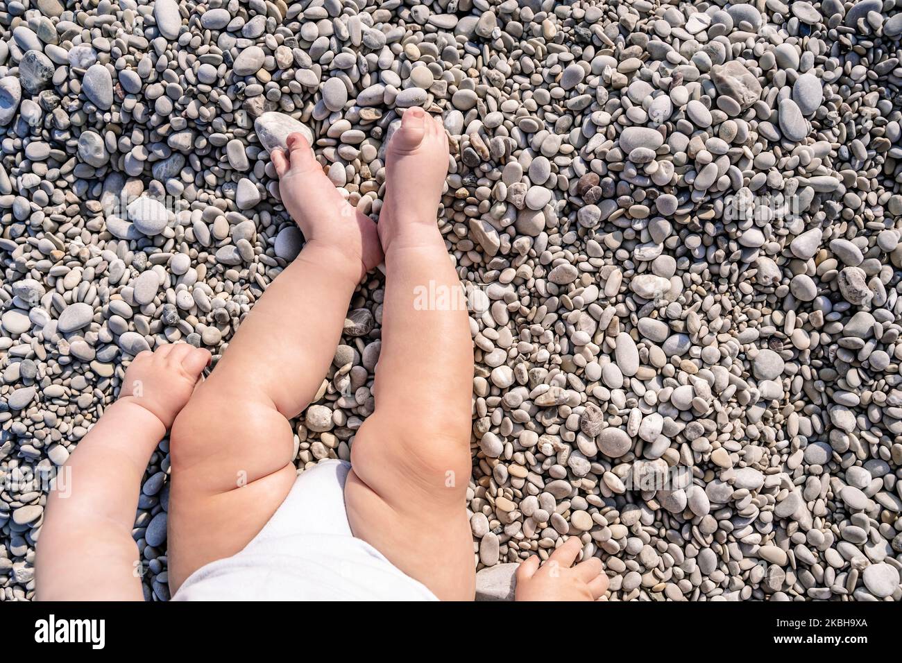Baby feet on the beach under the sun. Baby sits on a pebble beach in front of the sea in the summer on vacation. Health and care for young children. High quality photo Stock Photo