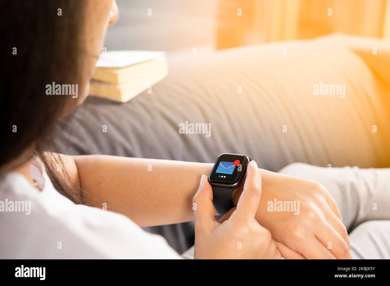 Unread email icon on smartwatch screen. Over shoulder view of woman touching her wearable technology, checking messages. Relaxing on the couch. Stock Photo