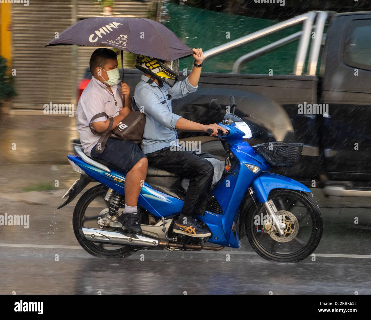 SAMUT PRAKAN, THAILAND, SEP 21 2022, A schoolboy rides a motorbike in the rain and holds an umbrella over himself and the driver Stock Photo