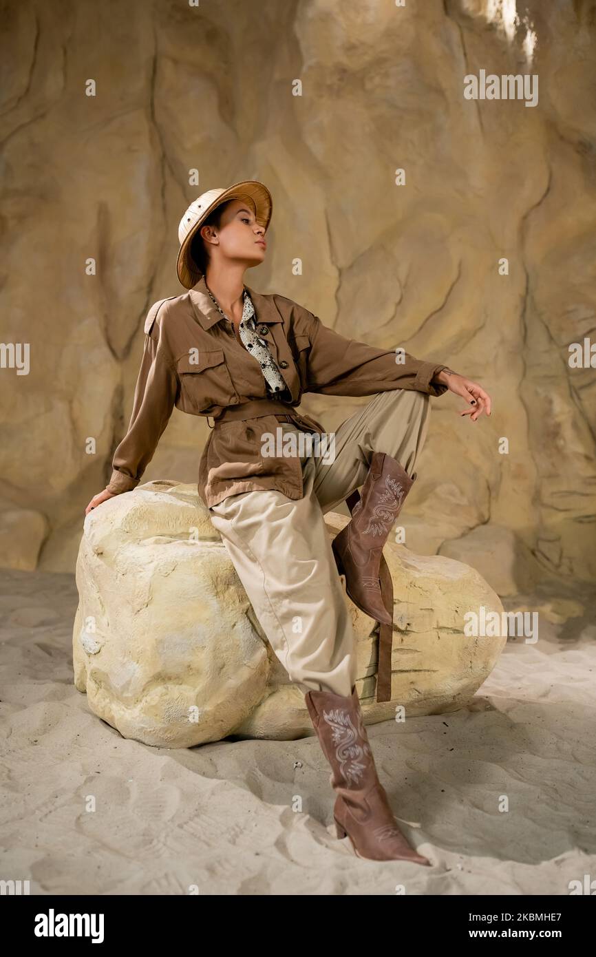 archaeologist in safari hat and brown jacket sitting on stone near rock in desert,stock image Stock Photo