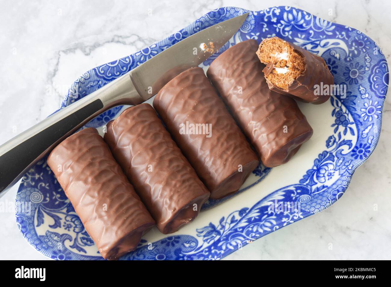 Individual chocolate sponge rolls on a patterned blue and white dish with a serving knife. On a white marble background Stock Photo