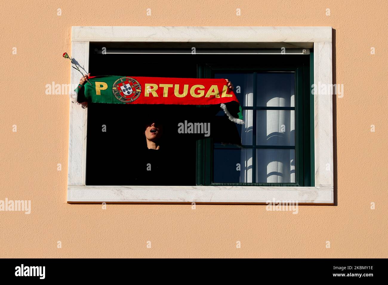 A woman holds a Portugals scarf and a red carnation (symbol of the 25 April 1974 Portuguese Revolution) as people sing the Portuguese revolution anthem 'Grandola Vila Morena' marking the 46th Anniversary of 25th April Revolution, in Lisbon, Portugal on April 25, 2020. The Portuguese celebrated the 46th anniversary of the Carnation Revolution today by singing at their windows, thereby circumventing the ban on gathering for traditional popular parades due to the coronavirus pandemic. (Photo by Pedro FiÃºza/NurPhoto) Stock Photo