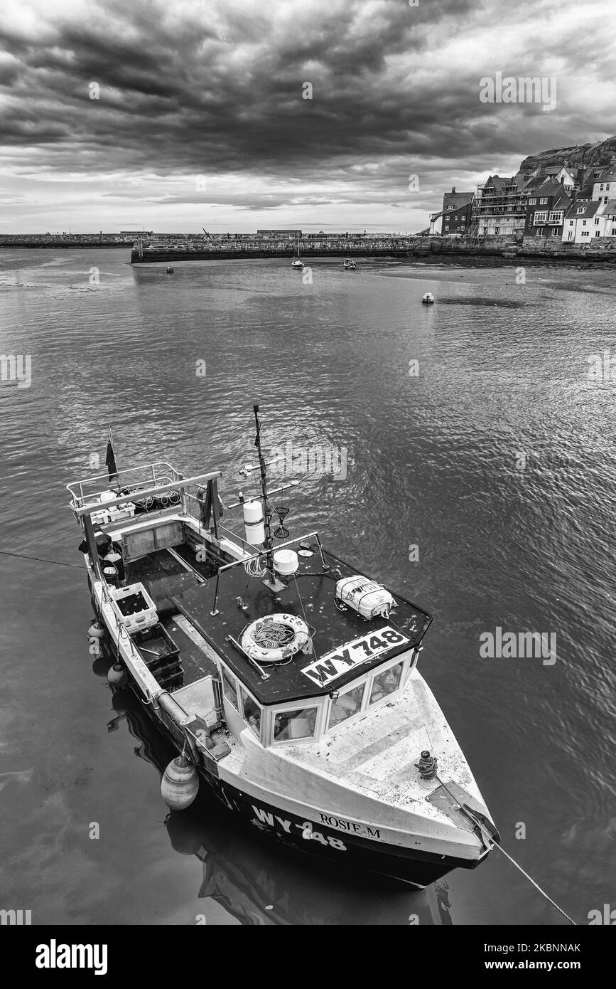 A fishing boat is moored in harbour. The harbour is beyond with houses by a cliff and a pier. A cloudy sky is overhead. Stock Photo