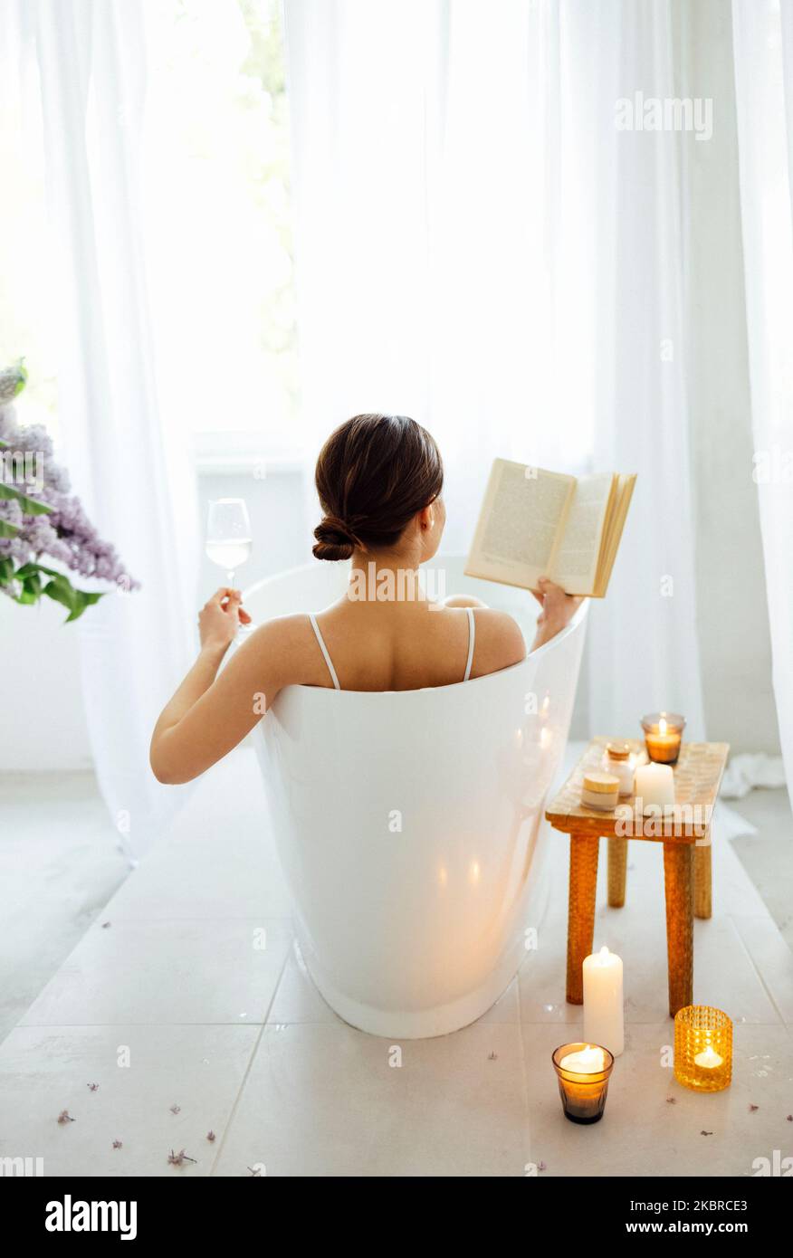 Back view of the young brunette woman reading the book and drinking white wine in bath with foam and candles Stock Photo