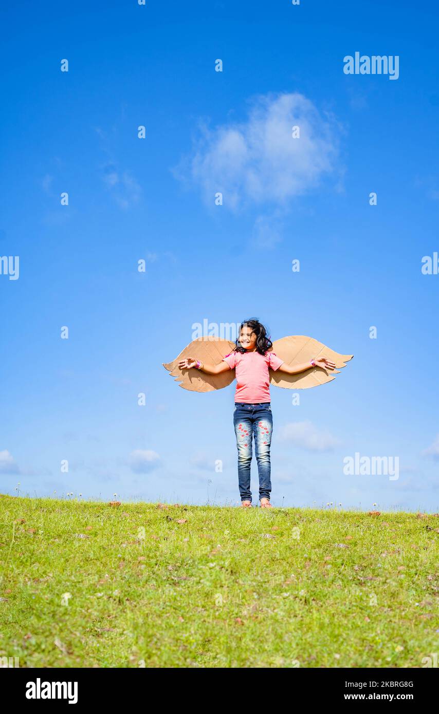 vertical shot, happy excited girl kid with artificial wings dancing like a bird on top hill in green meadow against blue sky - concept of freedom Stock Photo