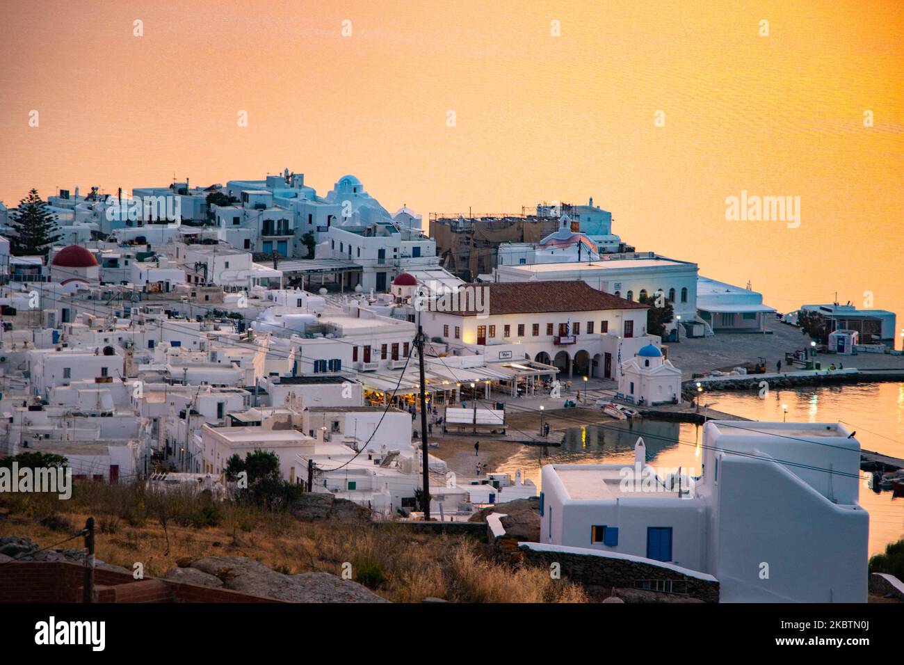 Dreamy sunset over Mykonos Town and the little harbor during the golden hour before dusk time with the sky shifting red, orange and warm colors as the sun goes down behind the Aegean Sea over Mykonos town or Chora, at Myconos Island in Cyclades, Aegean Sea in Greece. The famous Mediterranean Greek island is nicknamed as The Island of the Winds with whitewashed traditional buildings like windmills or little church. Mykonos is popular island for celebrities and tourists who want to party in the vibrant nightlife, the island is also considered gay friendly. Greek government relaunched the summer  Stock Photo