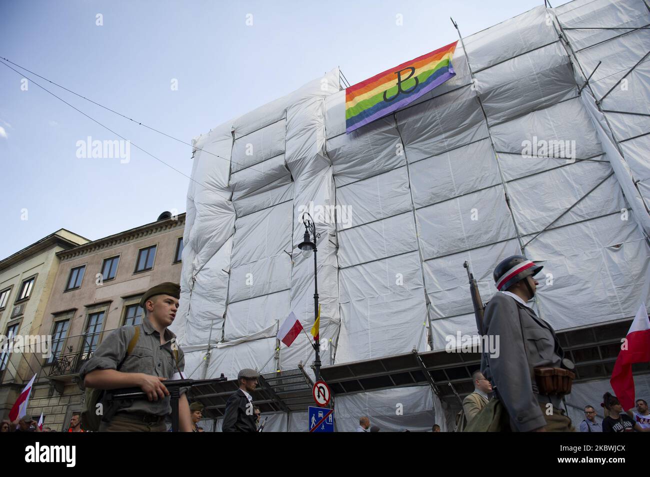 Rehearsal group members dressed as Polish underground soldiers walk by a rainbow flag with the Warsaw Uprising symbol (Powstanie Warszawskie), which has been hung as part of a counter demonstration during the 76th Warsaw uprising anniversary on August 1, 2020 in Warsaw, Poland. Each year, on the 1st of August at 5:00PM Varsovians take the streets to pay homage to the fallen soldiers of the Warsaw uprising that fought against the Nazi occupier during WWII. The rising was part of Operation Tempest that took place in 1944. (Photo by Aleksander Kalka/NurPhoto) Stock Photo
