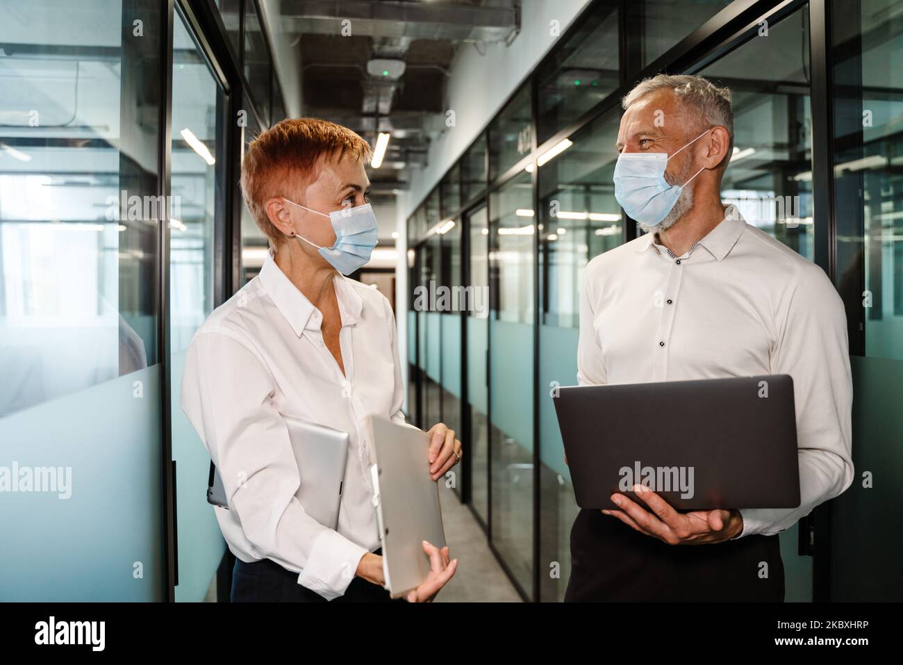 Two white colleagues in face masks talking while working in office Stock Photo