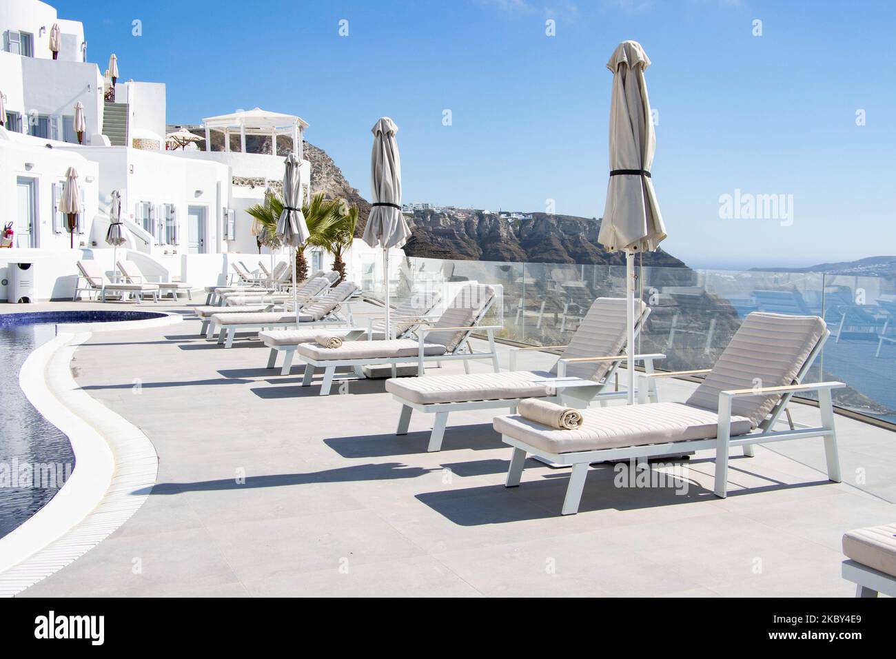 Swimming pool in a 5-star hotel resort with sun deck chairs and towel looking over a beautiful view on the Caldera over the Aegean sea with traditional whitewashed Greek Mediterranean terraced architecture of houses and resorts. Santorini Island in the Cyclades, Greece during a hot sunny summer day. Santorini is usually overcrowded as it is famous for its sunset attracting mostly couples from all over the world for the magical sunset view over the volcano, but this year due to the Coronavirus Covid-19 pandemic there are less than usual arrivals, reducing also the income of the GDP of the count Stock Photo