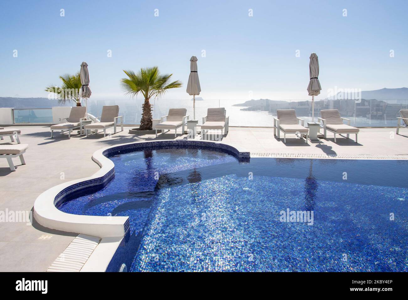 Swimming pool in a 5-star hotel resort with sun deck chairs and towel looking over a beautiful view on the Caldera over the Aegean sea with traditional whitewashed Greek Mediterranean terraced architecture of houses and resorts. Santorini Island in the Cyclades, Greece during a hot sunny summer day. Santorini is usually overcrowded as it is famous for its sunset attracting mostly couples from all over the world for the magical sunset view over the volcano, but this year due to the Coronavirus Covid-19 pandemic there are less than usual arrivals, reducing also the income of the GDP of the count Stock Photo