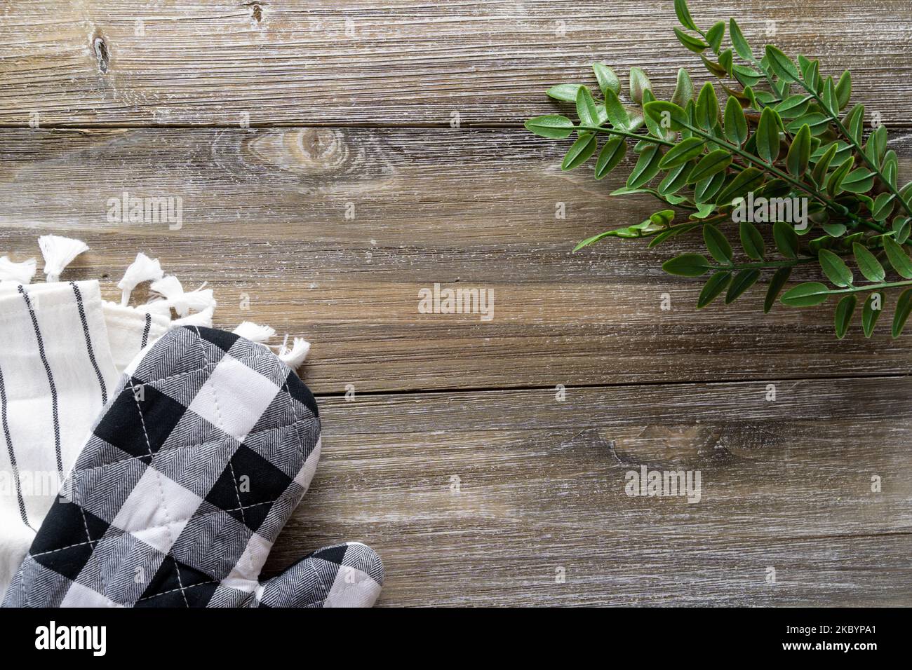 Baking concept flat lay, with black and white oven mitt, towel, and green leaves on a wood background Stock Photo