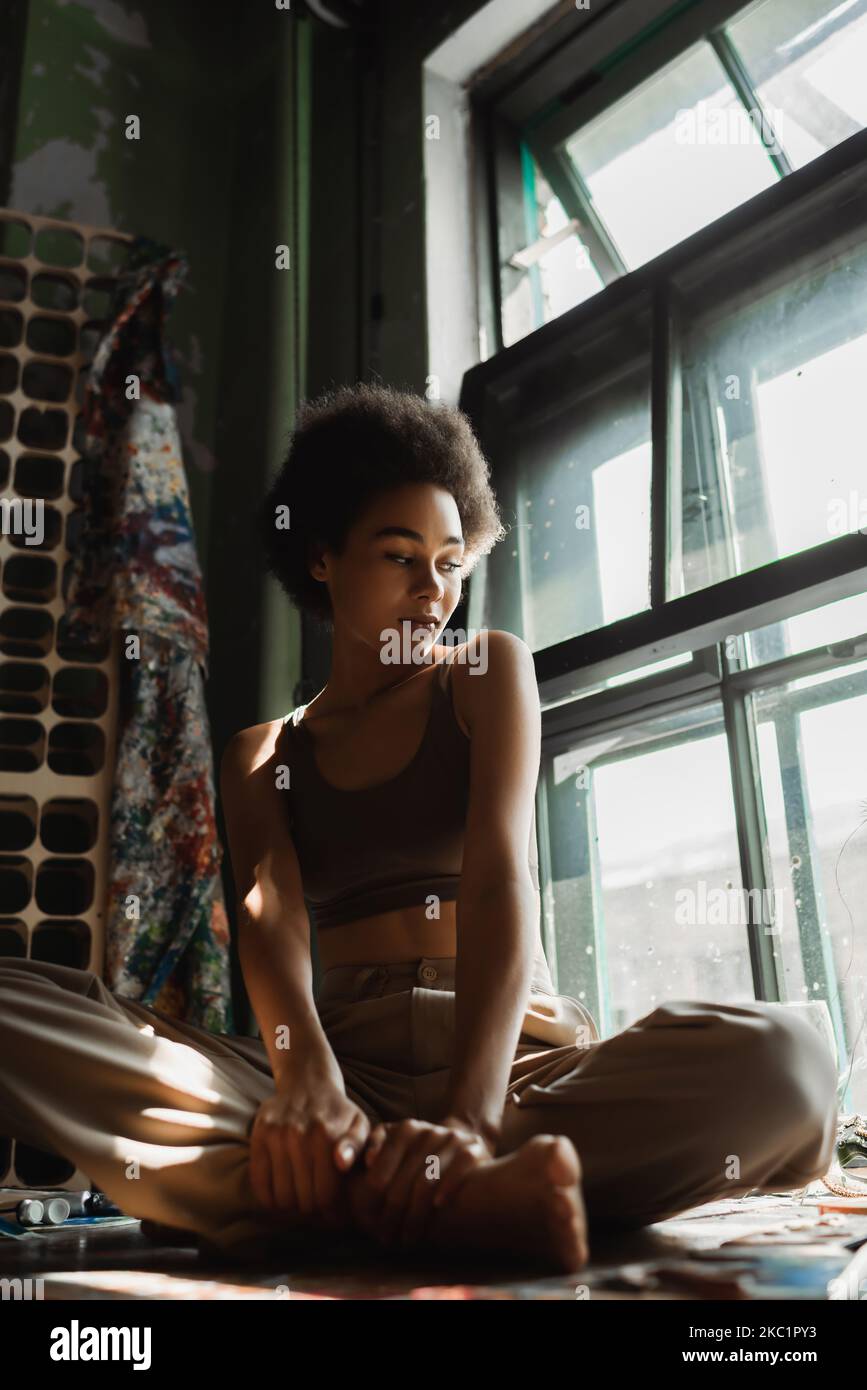 pensive african american woman sitting on windowsill with crossed legs ...