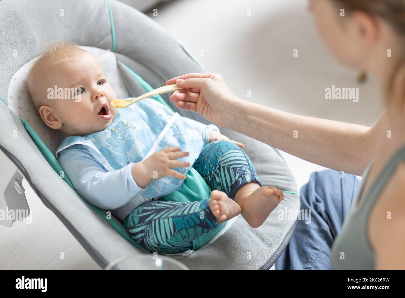 Mother spoon feeding her baby boy child in baby chair with fruit puree. Baby solid food introduction concept Stock Photo
