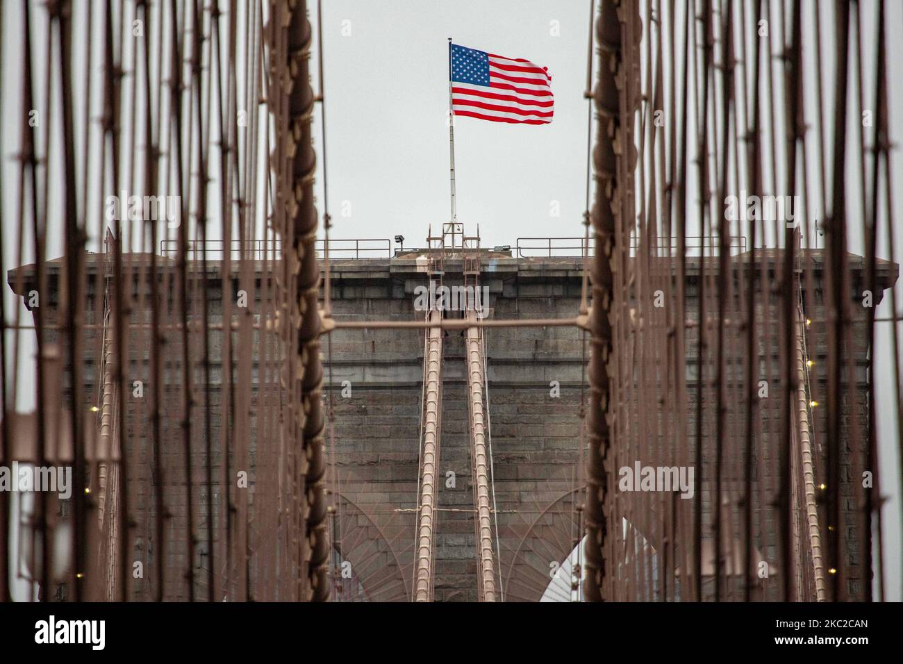 The US flag on the tower of the Brooklyn Bridge in New York City in the United States as seen during a cloudy day with tourists and locals on it. The famous bridge, a landmark for NYC and the United States of America is a hybrid cable stayed suspension bridge spanning the East River between the boroughs of Manhattan and Brooklyn. The historical NY landmark bridge was built between 1869 and 1883. New York, USA on February 13, 2020 (Photo by Nicolas Economou/NurPhoto) Stock Photo