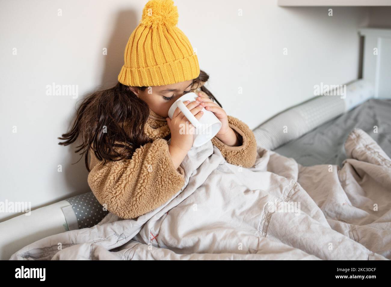 Beautiful little girl warms in blanket in cold winter time. Child in warm clothes sitting in a bed drinking hot tea at home. Cold home. No heating. Stock Photo