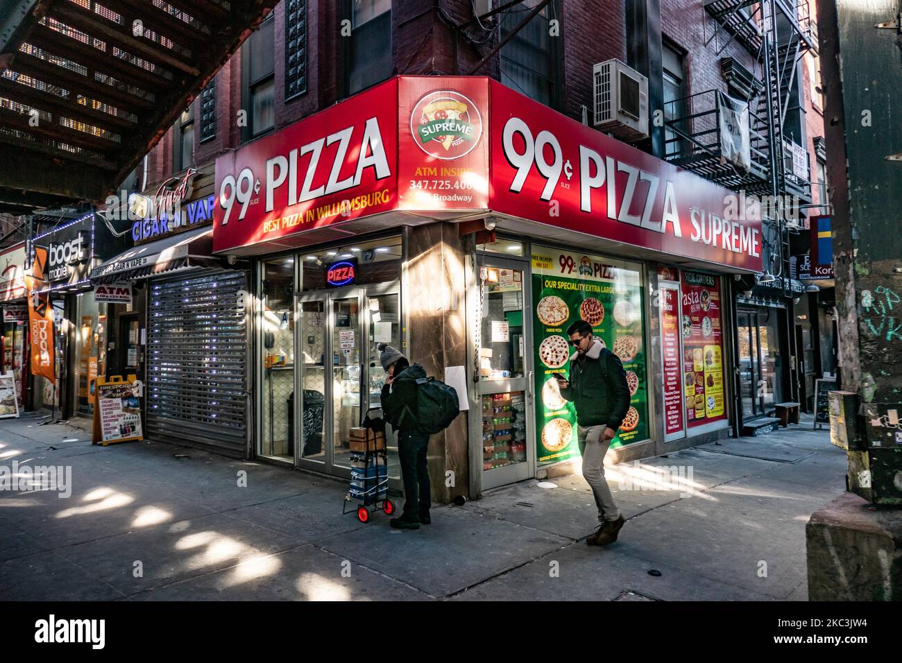 A man passing in front of 99c dolar slice pizza holding his phone and walking. Daily life view of the road and street under the subway railroad air-bridge metal construction of M, J and Z lines near Marcy Ave station, with restaurants, shops, the famous dollar slice pizza, traffic with cars and trucks and with sidewalks in Broadway street in Brooklyn, New York City. Brooklyn is the most populous county in the state of New York and the second-most densely populated in the United States. Brooklyn, NY, USA on 13 February 2020. (Photo by Nicolas Economou/NurPhoto) Stock Photo