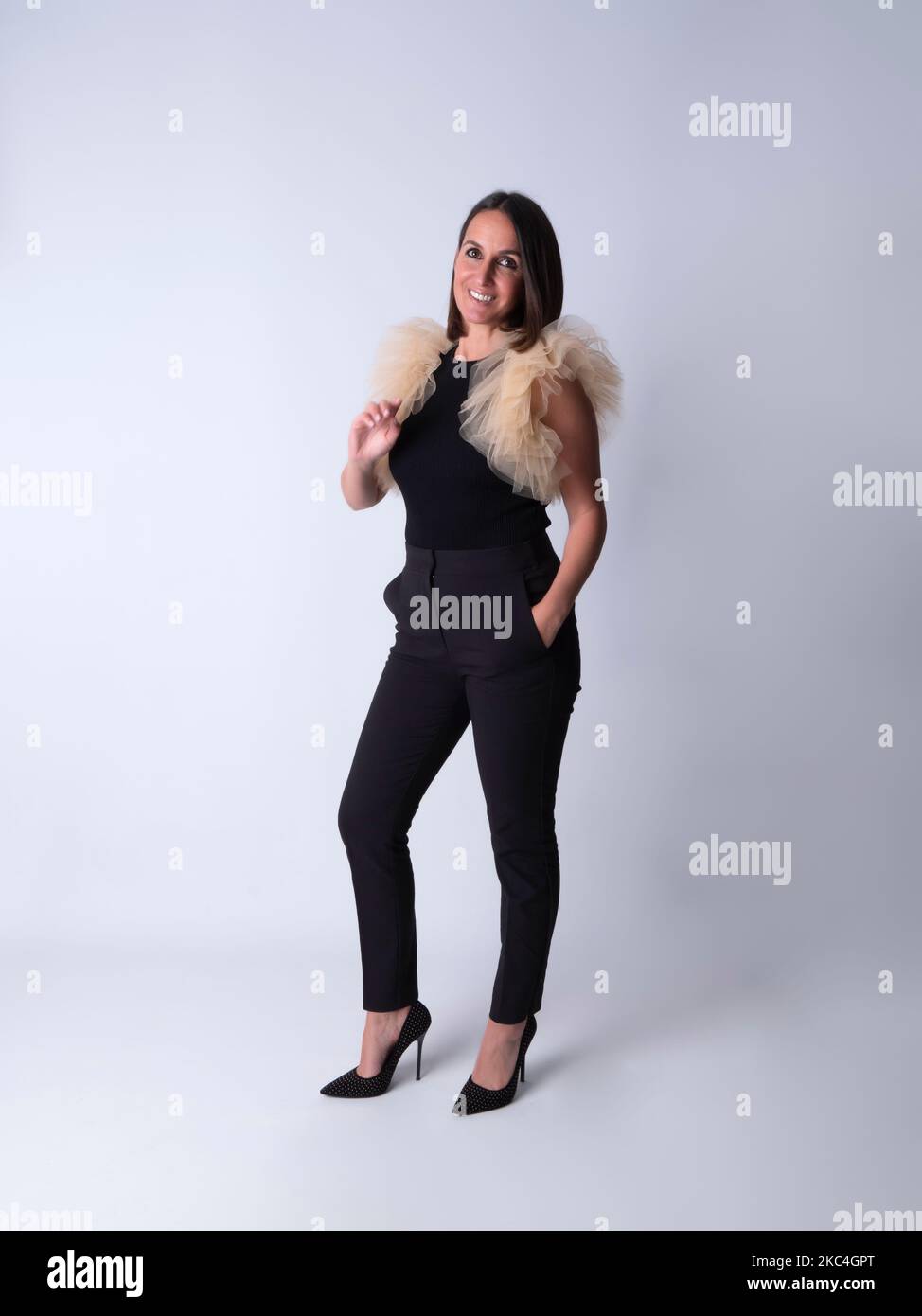 Full body glad adult brunette in stylish outfit touching chest and holding hand in pocket while standing against gray background and looking at camera Stock Photo