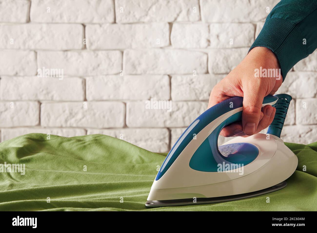 Man holding modern iron in hand and smooths wrinkled green shirt. Housework and laundry backgrounds. Copy space for text Stock Photo