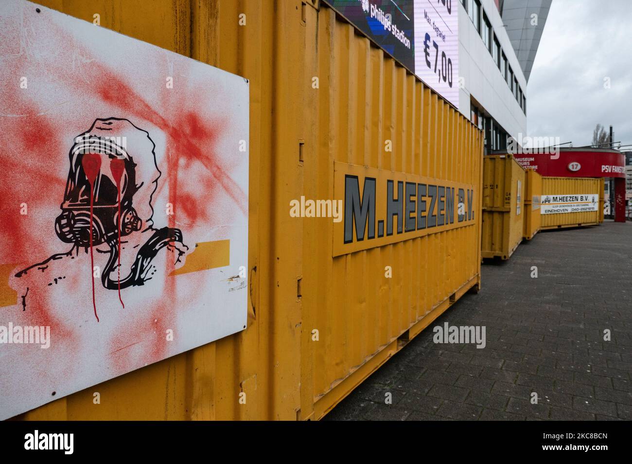 A sign of a man with chemical protection mask as seen vandalized with graffiti on the containers. The Philips Stadion, PSV club stadium in the Dutch city of Eindhoven is barricaded with shipping containers to protect the building, the museum, fanstore, multiple gates, trackside and supporters home side from riots and looting as happened previously in the city. The fortification and sealing of the front side of the stadium took place after the club received some threats according to the local media. Eindhoven faced some violent clashes during an anti-lockdown / anti-curfew protest that turned i Stock Photo