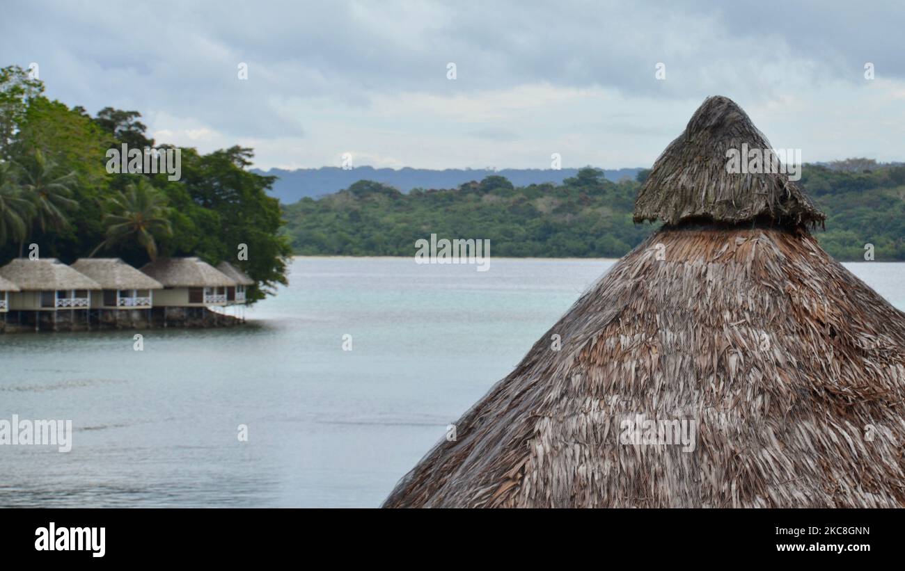 Roof cone of a traditional South Pacific Melanesian thatched building overlooks a lagoon with luxury overwater bungalows on the other side in Vanuatu Stock Photo