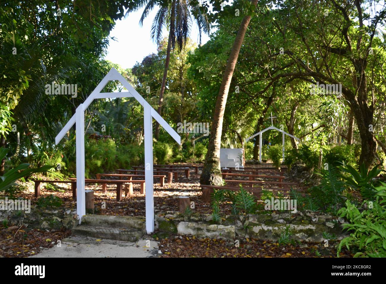 Outdoor chapel or church with entrance arch, pews, alter and religious cross amongst the lush foliage and palm trees in dappled sunlight in Vanuatu Stock Photo