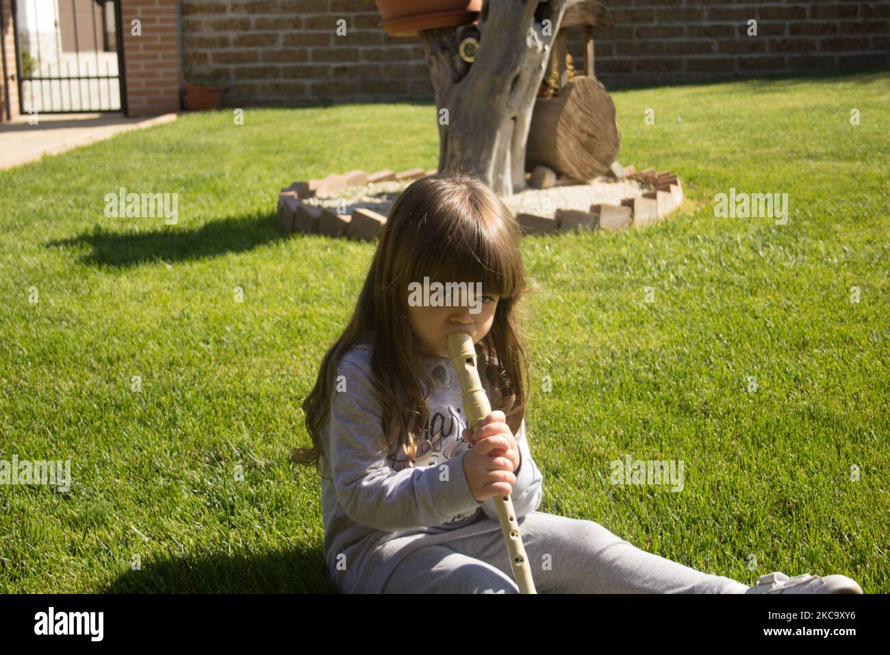Image of an adorable little brunette girl sitting on a green grass while learning to play the flute. Stock Photo