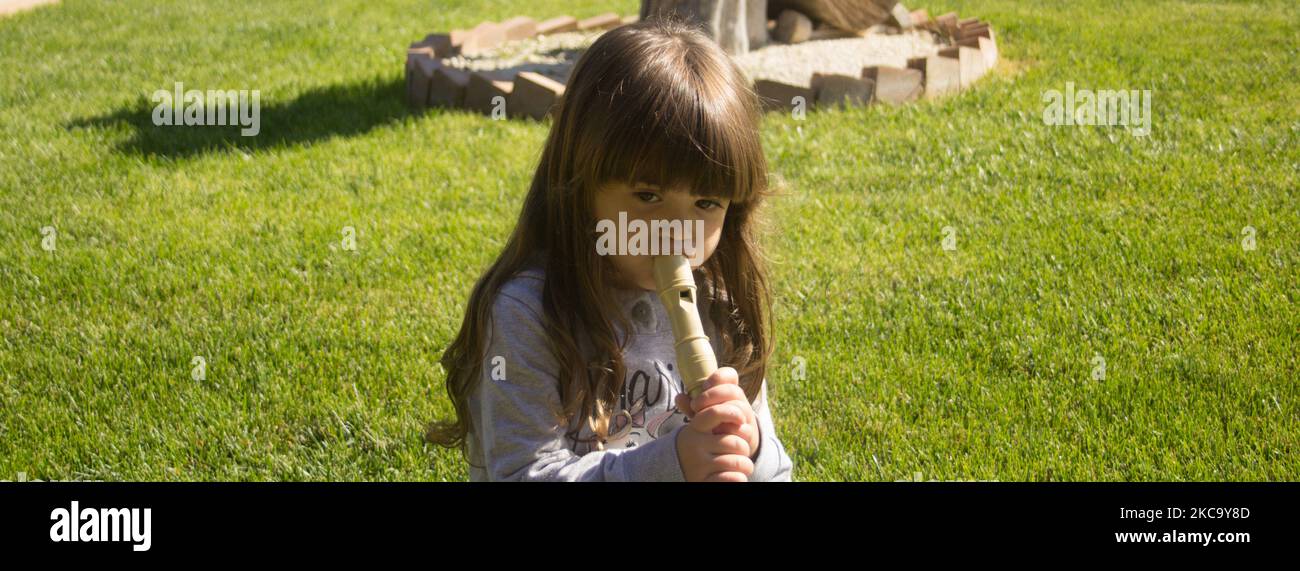 Image of an adorable little brunette girl sitting on a green grass while learning to play the flute. Horizontal banner Stock Photo