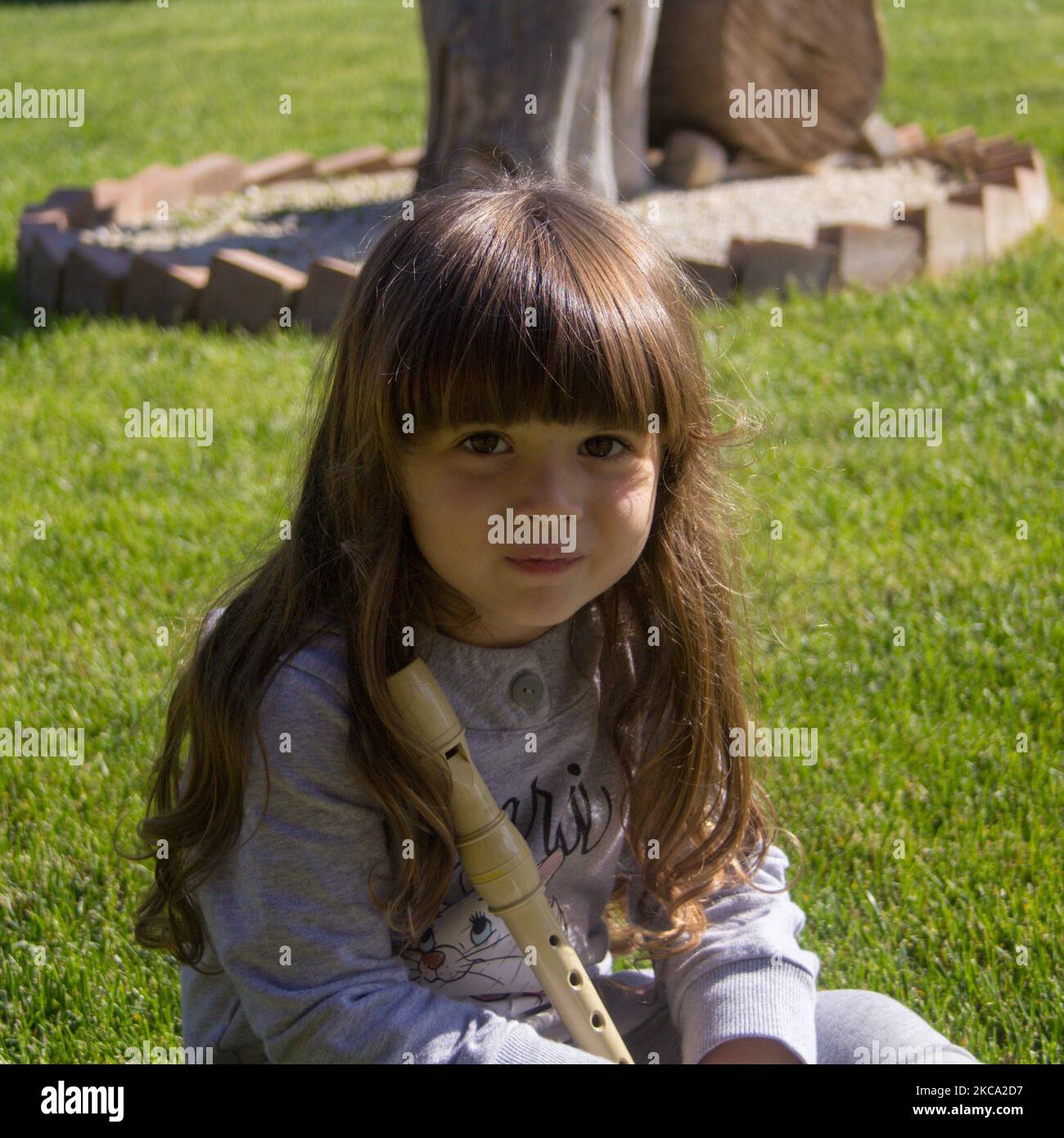 Image of an adorable smiling little girl posing sitting on a lawn holding a flute. Educational games for children to play outdoors Stock Photo