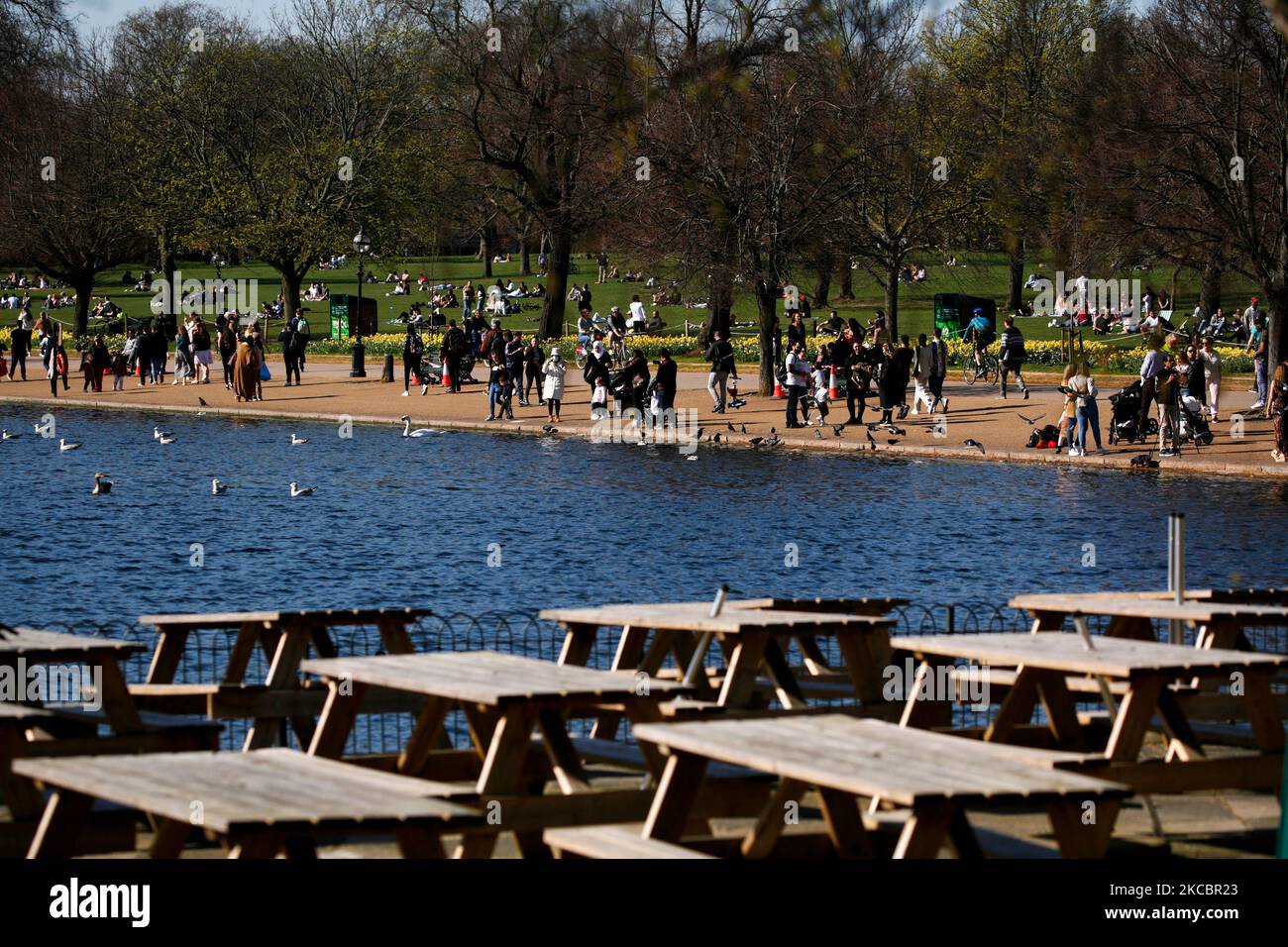 People walk in warm spring sunshine as empty tables sit out in the temporarily-closed outdoor seating area of a cafe beside the Serpentine lake in Hyde Park in London, England, on March 29, 2021. Lockdown restrictions were eased across England today, with the 'stay home' rule ended, groups of up to six allowed to meet outside, outdoor sports restarted and weddings with up to six people attending permitted to go ahead. Shops, hospitality and leisure businesses remain closed however, with the next relaxation of restrictions not due until April 12 at the earliest. Today's easing has meanwhile coi Stock Photo