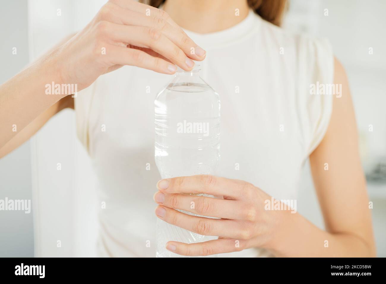 Close-up view of a young woman opening a plastic bottle of water standing by window at home, Health, people, food, sports, lifestyle and beauty Stock Photo