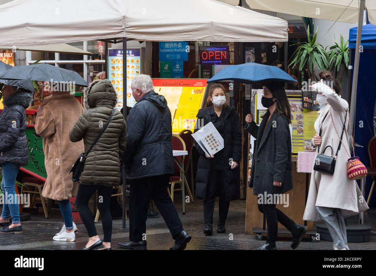 LONDON, UNITED KINGDOM - MAY 17, 2021: A waiter stands with a menu outside a restaurant in Chinatown while pedestrians shetlter from the rain as England moves to Step 3 in easing of coronavirus restrictions, on 17 May, 2021 in London, England. From today indoor hospitality and entertainment venues are allowed to re-open, six people or two households can meet indoors and international travel without need for quarantine resumes to 12 green list countries, however there are concerns that the surge in cases of the Indian Covid variant may delay the fourth stage of easing lockdown on June 21. (Phot Stock Photo