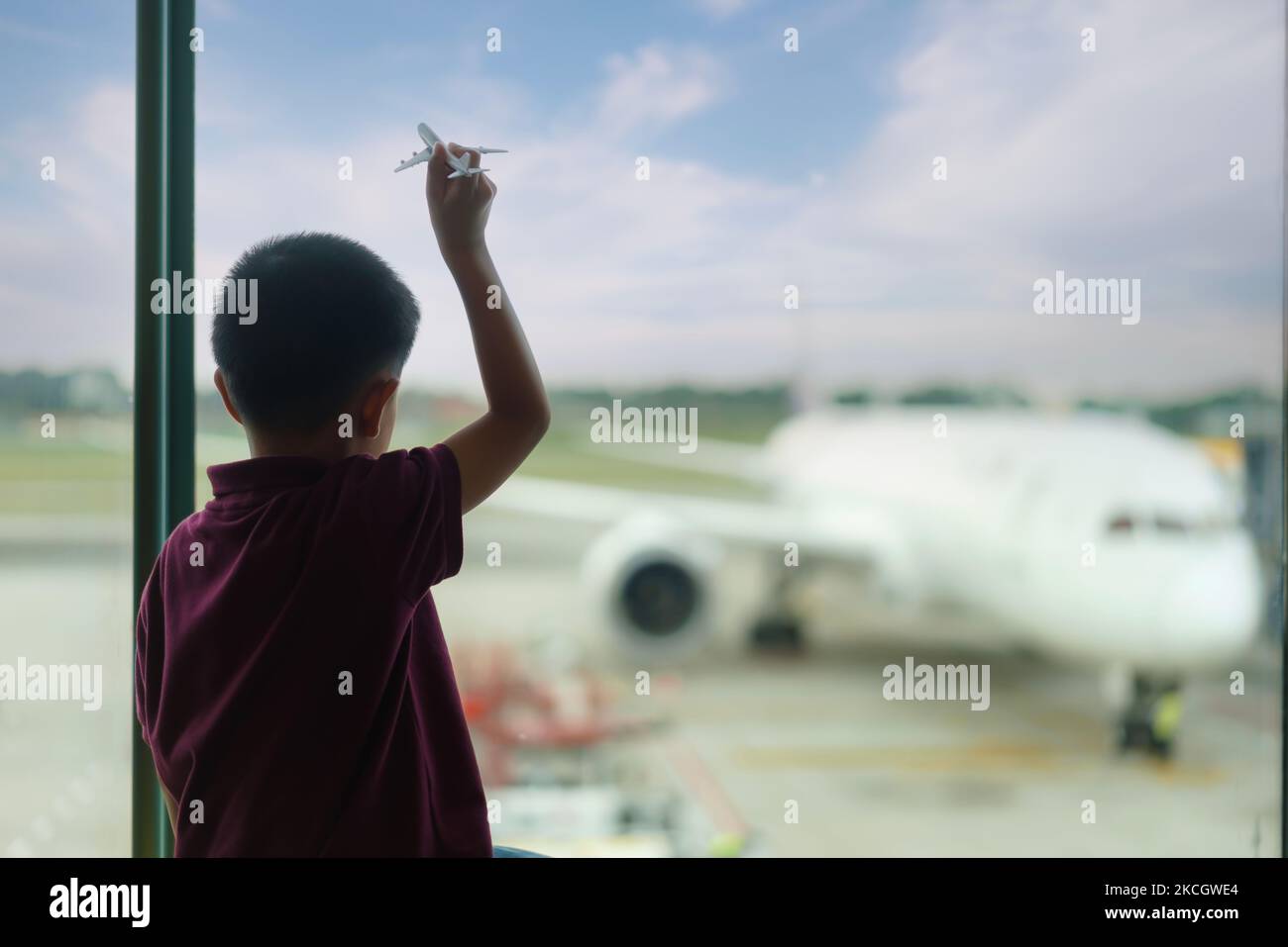 Asian Boy with airplane model at empty airport terminal waiting for departure looking out the window. Child in T-shirt and shorts stands at lounge wai Stock Photo