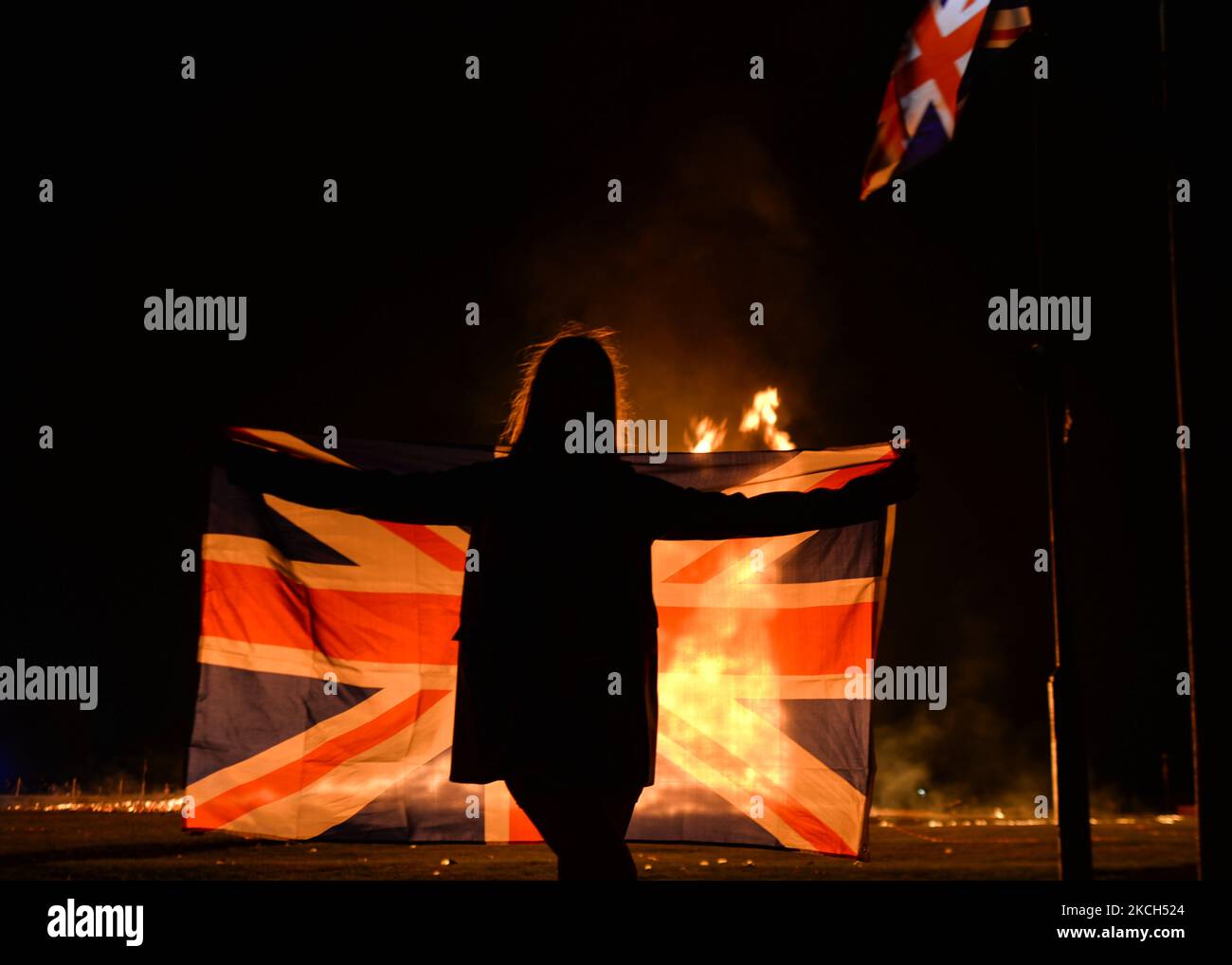 A person holding a Union Jack seen in front of a large bonfire during the Eleventh Night marking the start of the unionist Twelfth celebrations, in Craigy Hill, Larne. Tonight, large bonfires are lit in many Protestant loyalist neighbourhoods of Northern Ireland. Bonfires were originally lit to celebrate the Glorious Revolution (1688) and victory of Protestant king William of Orange over Catholic king James II at the Battle of the Boyne (1690), which began the Protestant Ascendancy in Ireland. On Monday, 12 July 2021, in Larne, County Antrim, Northern Ireland (Photo by Artur Widak/NurPhoto) Stock Photo
