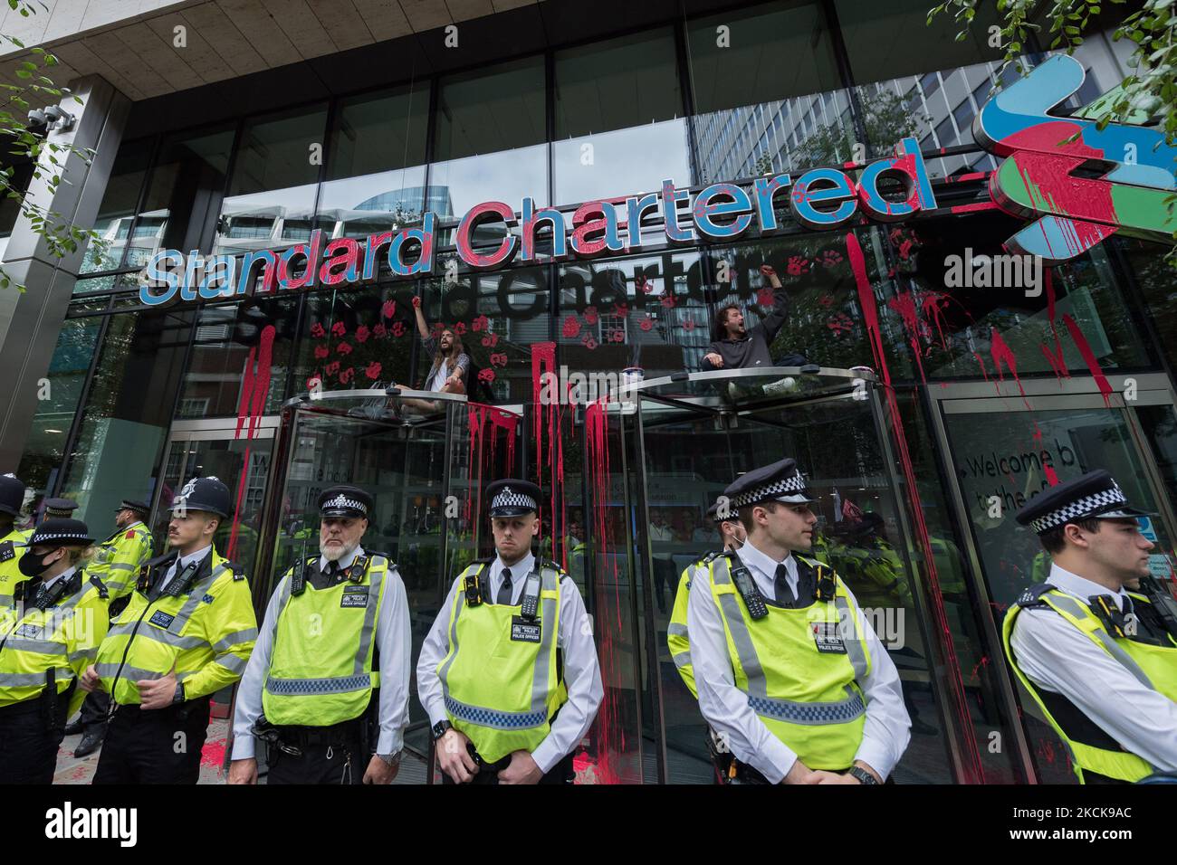 LONDON, UNITED KINGDOM - AUGUST 27, 2021: Environmental activists from Extinction Rebellion sit on top of the entrance to the Standard Chartered which has red paint spilled over the windows during a protest against the companies and institutions that are financing, insuring and enabling major fossil fuel projects and extraction of resources in the developing countries of the Global South, demanding change to the colonial system that drives the crises of climate and racism on 27 August 2021 in London, England. Extinction Rebellion activists target the City of London during the two week 'Impossi Stock Photo